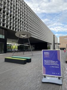 A photo of the Belval campus of the University of Luxembourg, with a sculpture of a large disk on tripod legs and a conference poster in the foreground